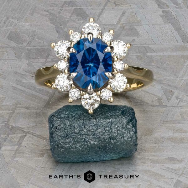 The "Angelica" ring in 14k white and yellow gold with 2.30-carat Montana sapphire The "Angelica" ring, crafted in 14k white and yellow gold with a 2.30-carat Montana sapphire surrounded by diamonds, is displayed on a rough blue stone. The Earth's Treasury logo appears below.