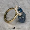 The "Angelica" ring in 14k white and yellow gold with a 2.30-carat Montana sapphire is balanced on a rough blue gem atop a textured gray surface, with the "Earth's Treasury" logo visible at the bottom.