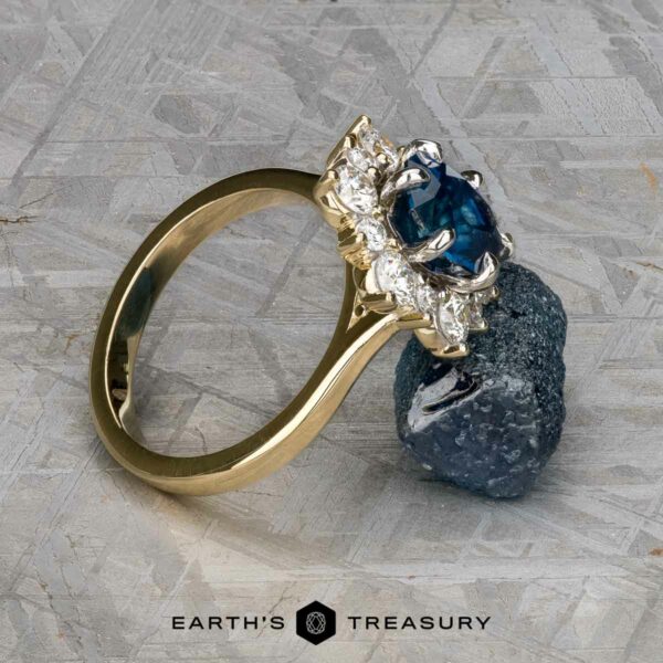 The "Angelica" ring in 14k white and yellow gold with 2.30-carat Montana sapphire The "Angelica" ring in 14k white and yellow gold with a 2.30-carat Montana sapphire is balanced on a rough blue gem atop a textured gray surface, with the "Earth's Treasury" logo visible at the bottom.
