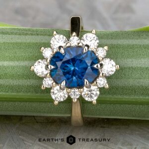 The "Angelica" ring in 14k white and yellow gold features a 2.30-carat Montana sapphire center surrounded by ten clear diamonds, displayed on a green-striped surface.