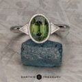 The "Ceres" ring in 14K white gold, featuring a 2.52-carat Madagascar sapphire accented by two triangular white side stones, is displayed upright on a rough blue-green rock.
