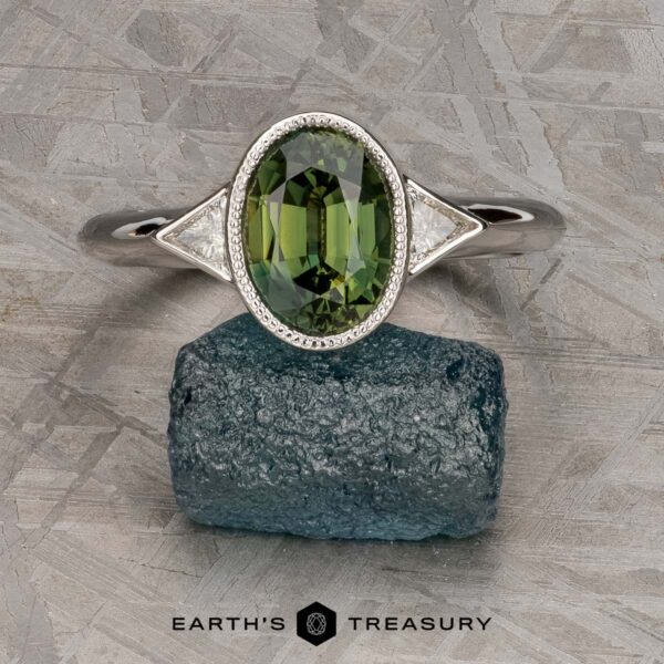 The "Ceres" ring in 14K white gold with 2.52-carat Madagascar sapphire The "Ceres" ring in 14K white gold, featuring a 2.52-carat Madagascar sapphire accented by two triangular white side stones, is displayed upright on a rough blue-green rock.