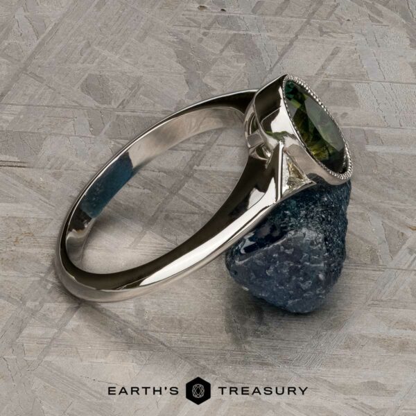 The "Ceres" ring in 14K white gold with 2.52-carat Madagascar sapphire The "Ceres" ring in 14K white gold with a 2.52-carat Madagascar sapphire rests on a rough blue stone; logo and text "EARTH'S TREASURY" appear at the bottom.