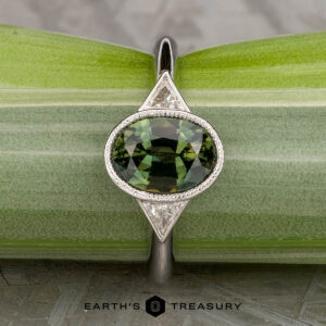 The "Ceres" ring in 14K white gold features a 2.52-carat Madagascar sapphire accented by two triangular clear stones, displayed on a light green plant stem.