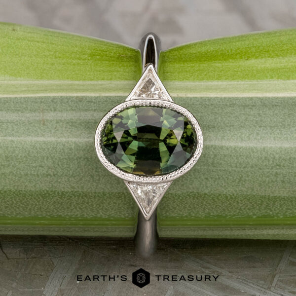 The "Ceres" ring in 14K white gold with 2.52-carat Madagascar sapphire The "Ceres" ring in 14K white gold features a 2.52-carat Madagascar sapphire accented by two triangular clear stones, displayed on a light green plant stem.