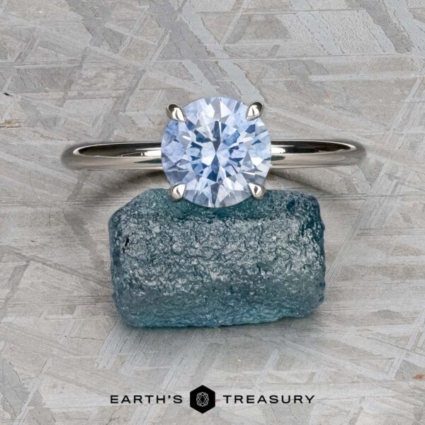 The "Tiara" ring in platinum with 2.00-carat Montana sapphire The "Tiara" ring in platinum with a 2.00-carat Montana sapphire is displayed on a rough blue gemstone, set against a textured gray background with the Earth’s Treasury logo at the bottom.