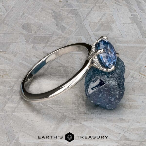 The "Tiara" ring in platinum with 2.00-carat Montana sapphire The "Tiara" ring in platinum, set with a 2.00-carat Montana sapphire, rests on a rough blue stone atop a textured gray surface. The "Earth's Treasury" logo is shown at the bottom.