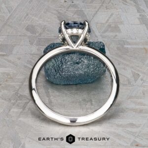 The "Tiara" ring in platinum features a 2.00-carat Montana sapphire accented by small clear stones, displayed upright on a textured gray stone, with the "Earth's Treasury" logo at the bottom.
