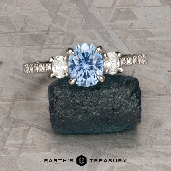 The Pave "Dryope" ring in platinum with 1.15-carat Montana sapphire The Pave "Dryope" ring in platinum features a 1.15-carat Montana sapphire flanked by two clear stones and a diamond-accented band, shown on dark rough stone with a textured background.