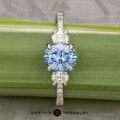 The Pave "Dryope" ring in platinum features a 1.15-carat round Montana sapphire center and two clear side stones, shown on a green stem with the "Earth's Treasury" logo below.