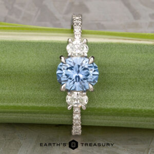 The Pave "Dryope" ring in platinum features a 1.15-carat round Montana sapphire center and two clear side stones, shown on a green stem with the "Earth's Treasury" logo below.