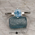 The "Bridget" ring in platinum, featuring a 1.20-carat Montana sapphire and two small rectangular side stones, is shown on a rough blue crystal with a textured background and "Earth's Treasury" branding.