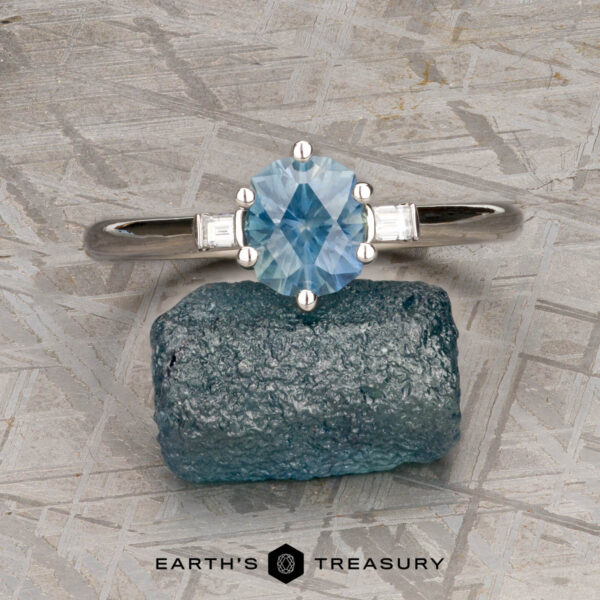 The "Bridget" ring in platinum with 1.20-carat Montana sapphire The "Bridget" ring in platinum, featuring a 1.20-carat Montana sapphire and two small rectangular side stones, is shown on a rough blue crystal with a textured background and "Earth's Treasury" branding.