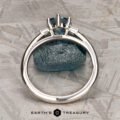 The "Bridget" ring in platinum, featuring a 1.20-carat Montana sapphire, is shown upright before a matching rough blue stone on a textured gray surface. The bottom text reads "Earth's Treasury.