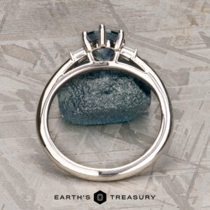 The "Bridget" ring in platinum, featuring a 1.20-carat Montana sapphire, is shown upright before a matching rough blue stone on a textured gray surface. The bottom text reads "Earth's Treasury.