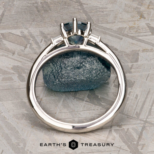 The "Bridget" ring in platinum with 1.20-carat Montana sapphire The "Bridget" ring in platinum, featuring a 1.20-carat Montana sapphire, is shown upright before a matching rough blue stone on a textured gray surface. The bottom text reads "Earth's Treasury.