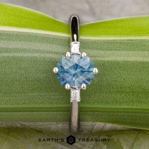 The "Bridget" ring in platinum features a 1.20-carat round Montana sapphire and two rectangular side stones, displayed on a green leaf background. The Earth's Treasury logo is visible at the bottom.