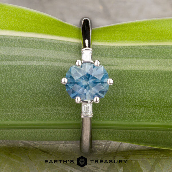 The "Bridget" ring in platinum with 1.20-carat Montana sapphire The "Bridget" ring in platinum features a 1.20-carat round Montana sapphire and two rectangular side stones, displayed on a green leaf background. The Earth's Treasury logo is visible at the bottom.
