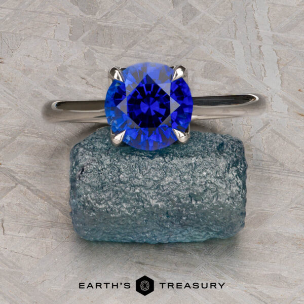 The Cathedral "Alyssum" ring in platinum with 1.83-carat Ceylon sapphire The Cathedral "Alyssum" platinum ring with a 1.83-carat Ceylon sapphire rests on rough blue crystal against a textured gray background, with "Earth's Treasury" at the bottom.