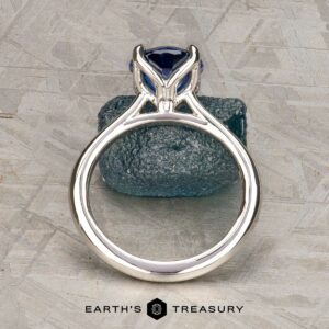 A platinum Cathedral "Alyssum" ring with a 1.83-carat Ceylon sapphire stands upright on a textured gray surface, exuding elegance. The "Earth's Treasury" logo appears at the bottom.