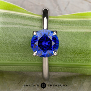 A platinum Cathedral "Alyssum" ring featuring a 1.83-carat Ceylon sapphire is displayed upright against a green leaf background. The "Earth's Treasury" logo appears at the bottom.