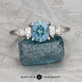 The "Tigrina" ring in platinum, featuring a 1.25-carat Montana sapphire and diamond accents, is displayed on a rough blue stone with a textured gray background and the “Earth’s Treasury” logo at the bottom.
