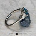 The "Tigrina" ring in platinum, featuring a 1.25-carat Montana sapphire and accent diamonds, rests on a textured blue mineral; “EARTH'S TREASURY” appears underneath.