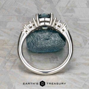 The "Tigrina" ring in platinum with a 1.25-carat Montana sapphire is displayed upright, highlighting its setting and band, against a blue stone background with the "Earth's Treasury" logo below.