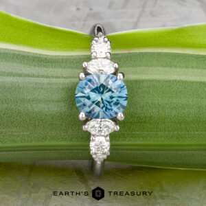 The "Tigrina" ring in platinum features a 1.25-carat round Montana sapphire center, accented by three clustered white stones on each side, shown against a green leaf background.