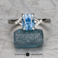The "Tango" ring in platinum, featuring a 2.03-carat Montana sapphire and accent diamonds, is displayed atop a rough blue stone on a gray patterned surface; the "Earth's Treasury" logo appears at the bottom.