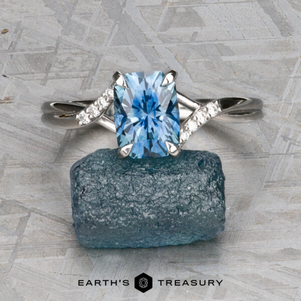 The "Tango" ring in platinum with 2.03-carat Montana sapphire The "Tango" ring in platinum, featuring a 2.03-carat Montana sapphire and accent diamonds, is displayed atop a rough blue stone on a gray patterned surface; the "Earth's Treasury" logo appears at the bottom.