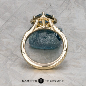 The "Yvette" ring in 14k yellow gold with a 1.64-carat Montana sapphire is shown upright on a rough blue stone, with the Earth's Treasury logo below.