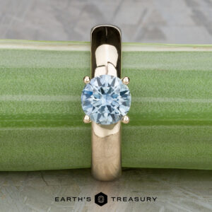 The "Davina" ring, crafted in 14k yellow gold with a 1.43-carat Montana sapphire, is displayed upright against a green bamboo stem. The "Earth's Treasury" logo appears below.