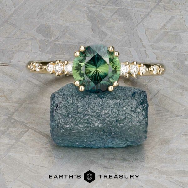 The "Blythe" ring in 14k yellow gold with 1.51-carat sapphire The "Blythe" ring in 14k yellow gold features a 1.51-carat green sapphire and small side diamonds, displayed on a rough blue-green crystal with the "Earth's Treasury" logo below.