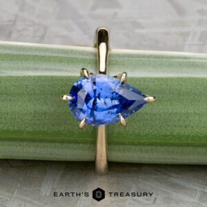 A gold Cathedral Solitaire ring, "Fisher, Matthew," with a pear-shaped blue gemstone is shown on a green background, with "Earth's Treasury" at the bottom.