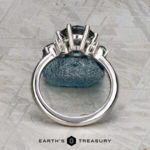 The "Xena" ring in platinum with a 2.27-carat Australian sapphire stands upright on a rough blue stone, photographed on a textured grey surface with the "Earth's Treasury" logo at the bottom.