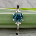 The "Xena" ring features a round blue-green 2.27-carat Australian sapphire with small diamonds in a platinum band, displayed on a green leaf, with the Earth's Treasury logo at the bottom.