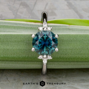 The "Xena" ring features a round blue-green 2.27-carat Australian sapphire with small diamonds in a platinum band, displayed on a green leaf, with the Earth's Treasury logo at the bottom.