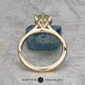 The "Carmen" ring in 14k yellow gold with a 1.29-carat Montana sapphire stands upright on a textured blue stone, set against a patterned gray background. The Earth's Treasury logo is visible below.