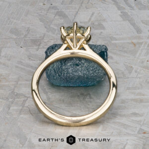 The "Carmen" ring in 14k yellow gold with a 1.29-carat Montana sapphire stands upright on a textured blue stone, set against a patterned gray background. The Earth's Treasury logo is visible below.