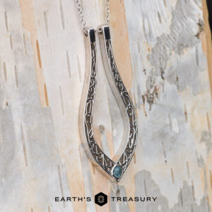 The "Nigella" ring keeper necklace in 14K white gold, featuring an intricate leaf design and a blue gemstone, hangs in front of a pale tree trunk with the text "EARTH'S TREASURY" below.