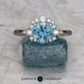 The "Veronica" ring in 14k white gold features a 1.53-carat Montana sapphire surrounded by small clear stones, displayed atop a rough blue mineral with the Earth's Treasury logo below.
