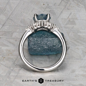 The "Veronica" ring in 14k white gold with a 1.53-carat Montana sapphire stands upright against a rough blue mineral on a textured gray background. The Earth’s Treasury logo appears at the bottom.