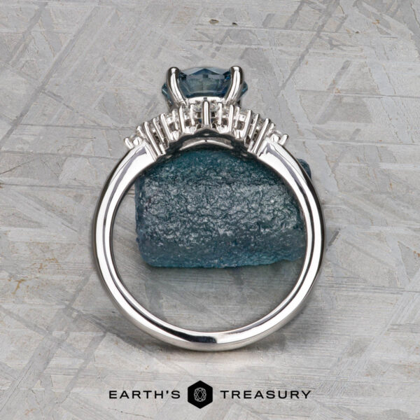 The "Veronica" ring in 14k white gold with 1.53-carat Montana sapphire The "Veronica" ring in 14k white gold with a 1.53-carat Montana sapphire stands upright against a rough blue mineral on a textured gray background. The Earth’s Treasury logo appears at the bottom.