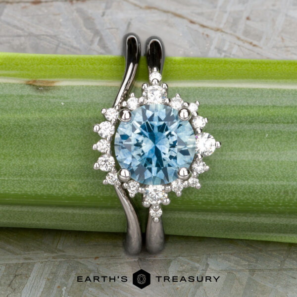 The "Veronica" in 14k white gold with 1.53-carat Montana sapphire, alongside the "Veronica" band in 14k white gold The "Veronica" ring in 14k white gold, featuring a 1.53-carat Montana sapphire and a halo of diamonds, is displayed with the matching "Veronica" band atop a green plant stalk. Logo at the bottom: Earth's Treasury.