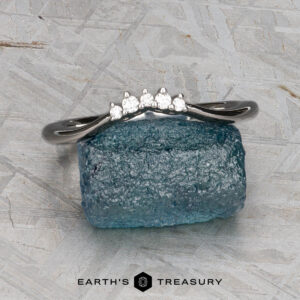 The "Veronica" band in 14k white gold, featuring five small round diamonds, rests on a rough blue gemstone against a textured gray background. The Earth’s Treasury logo appears at the bottom.