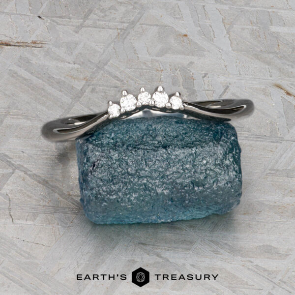 The "Veronica" band in 14k white gold The "Veronica" band in 14k white gold, featuring five small round diamonds, rests on a rough blue gemstone against a textured gray background. The Earth’s Treasury logo appears at the bottom.