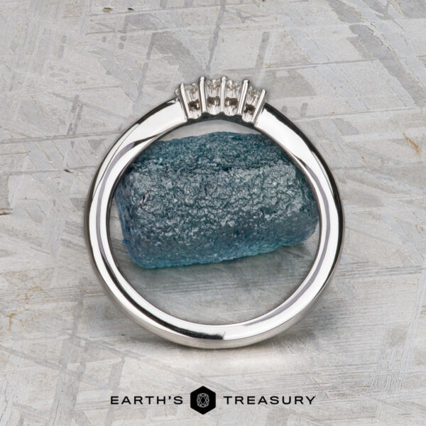 The "Veronica" band in 14k white gold The "Veronica" band in 14k white gold with four diamonds stands upright before a blue rough gemstone on a textured gray surface. The Earth’s Treasury logo appears at the bottom.