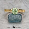 The "Maya" ring in 18k yellow gold, set with a 1.48-carat Australian sapphire, is displayed balanced on a raw blue-green crystal against a textured gray background.