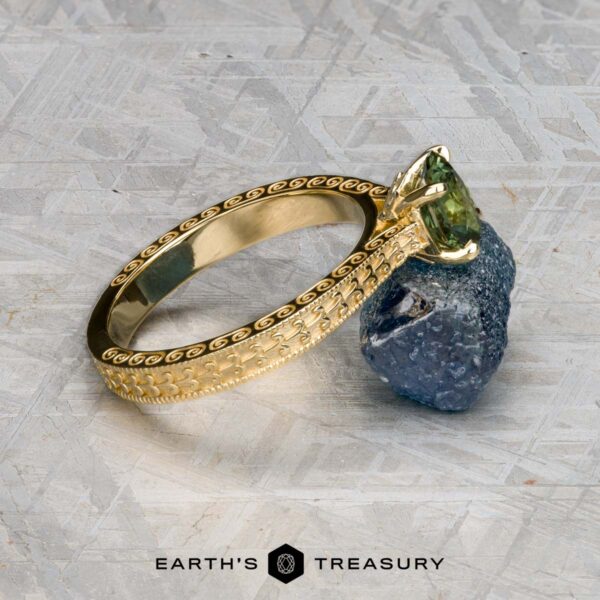 The "Maya" ring in 18k yellow gold with 1.48-carat Australian sapphire The "Maya" ring in 18k yellow gold, featuring a 1.48-carat Australian sapphire and intricate pattern, rests on a rough blue stone above a textured surface with the Earth's Treasury logo.